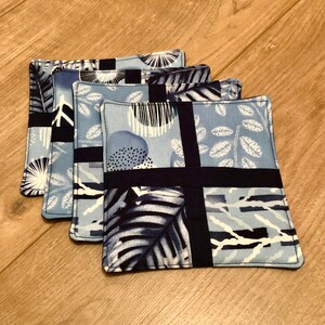 May include: Four blue and white fabric coasters with black trim. The coasters feature a patchwork design with different patterns, including leaves, stripes, and geometric shapes.