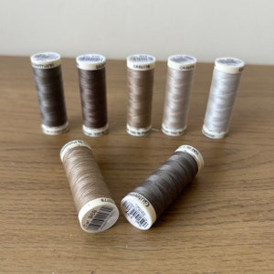 May include: A collection of ten spools of Gutermann thread in various shades of brown and grey. The spools are labelled with the Gutermann brand name and the colour number. The thread is 100% polyester.