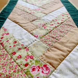 May include: A green and white patchwork table runner with a floral pattern. The runner is made of different fabrics with a variety of floral prints, including roses, small flowers, and a white fabric with a subtle pattern.