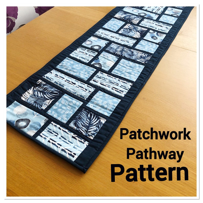 May include: A blue and white patchwork table runner with a geometric pattern. The runner is made of fabric squares with different designs, including leaves, circles, and stripes. The text "Patchwork Pathway Pattern" is printed below the runner.