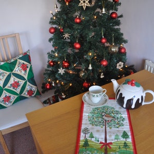 May include: A Christmas tree decorated with red ornaments, gold bells, and wooden stars. A teacup and teapot are on a table with a festive table runner featuring a bird and a tree.