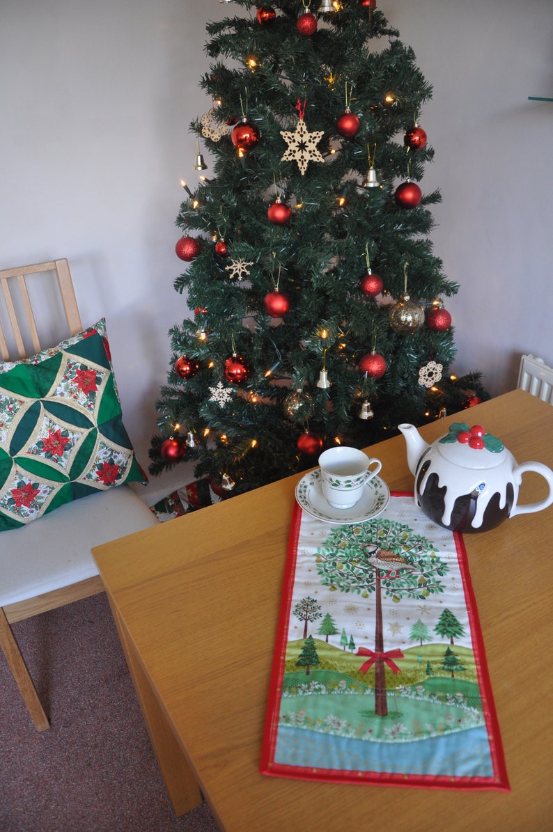 May include: A Christmas tree decorated with red ornaments, gold bells, and wooden stars. A teacup and teapot are on a table with a festive table runner featuring a bird and a tree.