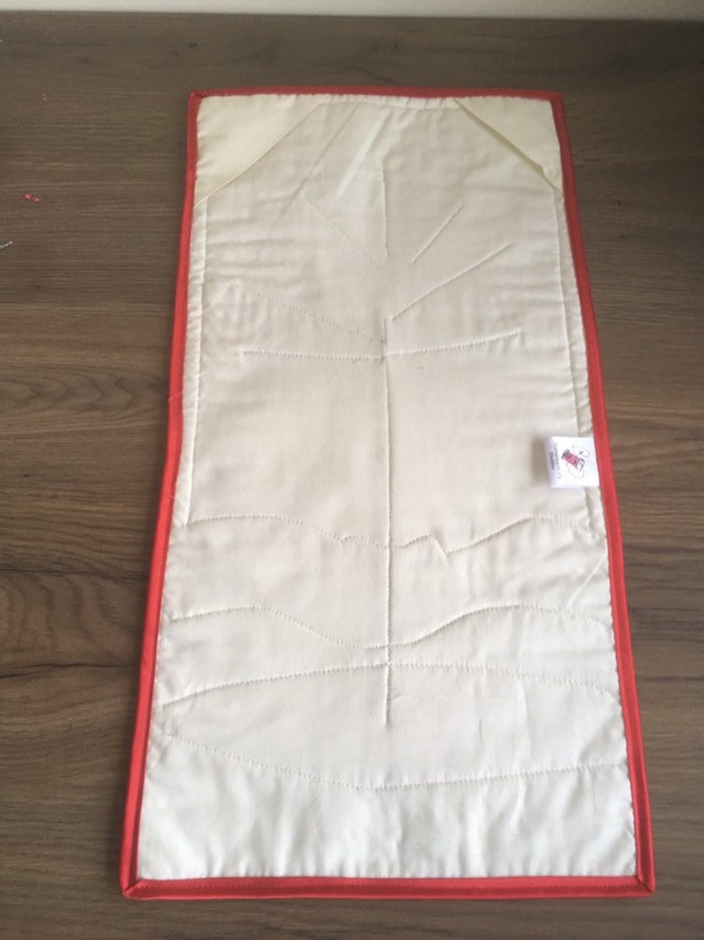 May include: A white quilted placemat with a red border. The placemat has a quilted design of a tree branch with leaves.