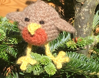 Könnte beinhalten: Ein brauner und roter gestrickter Rotkehlchen-Anhänger, der an einem grünen Weihnachtsbaum hängt.