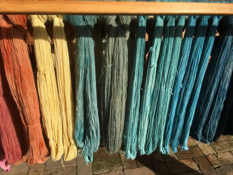 K&ouml;nnte beinhalten: Eine Pr&auml;sentation von gef&auml;rbtem Garn in verschiedenen Farben, darunter Rosa-, Orange-, Gelb-, Gr&uuml;n- und Blaut&ouml;ne. Das Garn h&auml;ngt an einem Holzgestell, wobei jedes Garnb&uuml;ndel oben gebunden ist. Das Bild ist gut beleuchtet und zeigt die Textur und Farbt&ouml;ne des Garns.