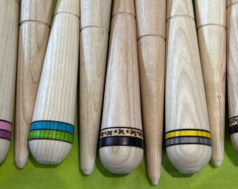 Könnte beinhalten: Ein Satz von 10 Stricknadeln aus Holz mit verschiedenen farbigen Bändern. Die Nadeln sind alle gleich groß und geformt, aber jede hat ein anderes farbiges Band um die Mitte. Die Bänder sind alle aus verschiedenen Materialien wie Leder, Stoff und Garn.