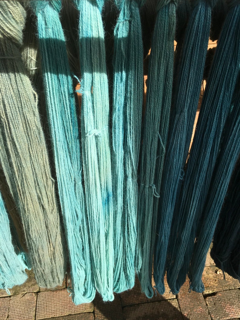 K&ouml;nnte beinhalten: Verschiedene Str&auml;nge gef&auml;rbter Wolle in Blau- und Gr&uuml;nt&ouml;nen, die vertikal h&auml;ngen. Die Farben reichen von hellem T&uuml;rkis bis zu tiefem Teal, mit sichtbarer Textur und Farbtonvariationen. Das Bild ist gut beleuchtet und zeigt die Naturfasern.