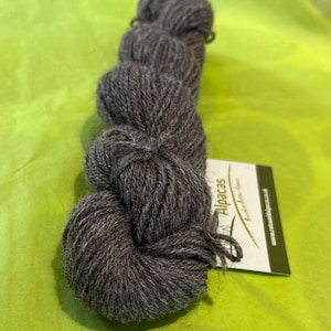 Puede incluir: Un ovillo de hilo de alpaca gris oscuro con una etiqueta blanca que dice "Alpacas" y "Super Soft Alpaca Yarn".