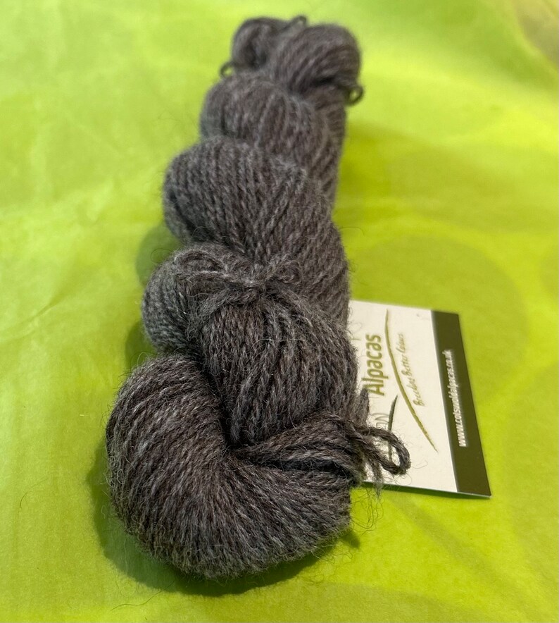 Puede incluir: Un ovillo de hilo de alpaca gris oscuro con una etiqueta blanca que dice "Alpacas" y "Super Soft Alpaca Yarn".
