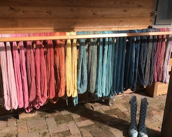 Puede incluir: Un estante de madera exhibe madejas de hilo colorido en una variedad de tonos, desde rosas y rojos hasta azules y grises. El hilo está colgado al aire libre, con un par de botas de agua azules con lunares en primer plano.