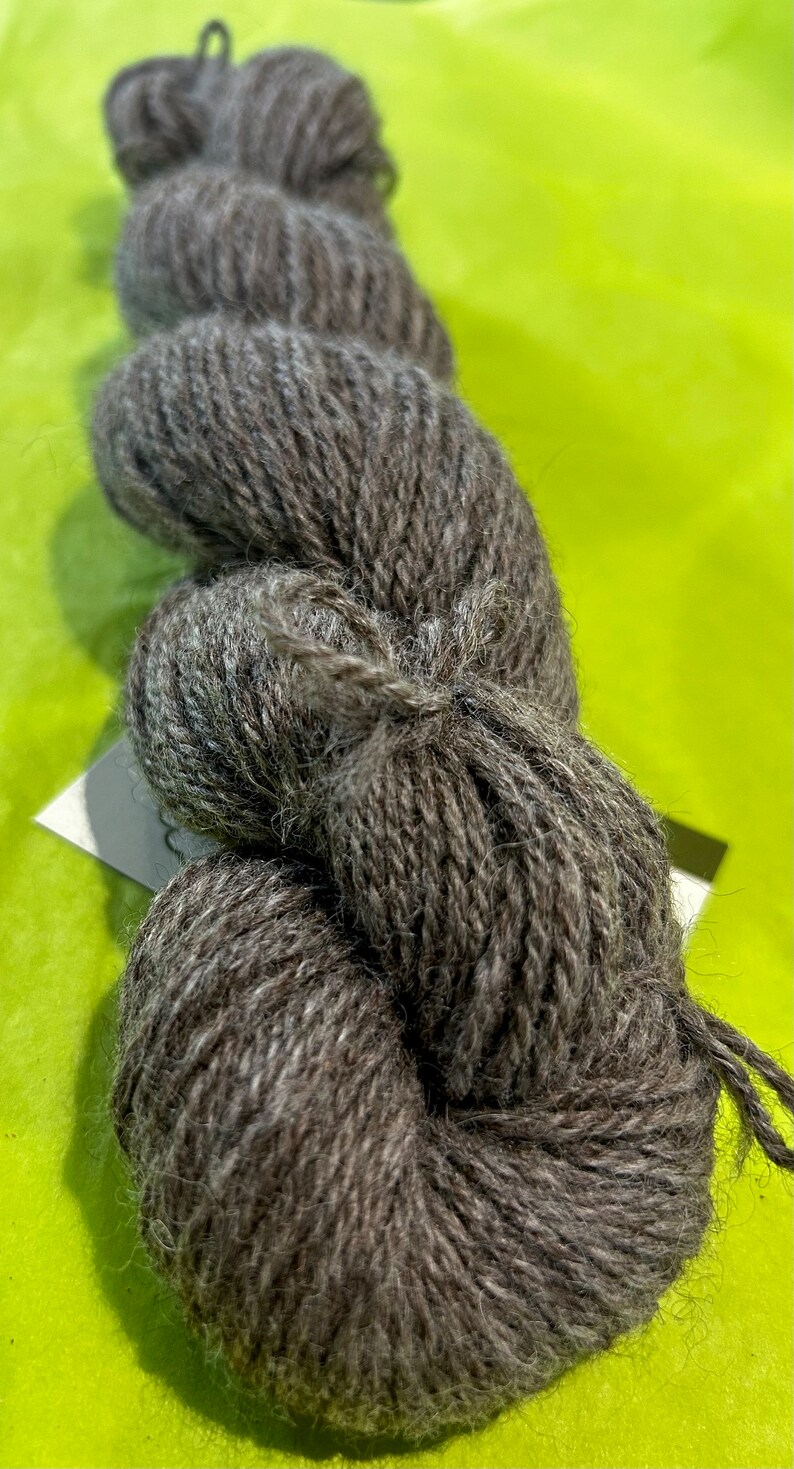 Puede incluir: Un ovillo de lana gris oscuro con una textura peluda. La lana est&aacute; enrollada en forma de bola y est&aacute; lista para tejer o hacer ganchillo.