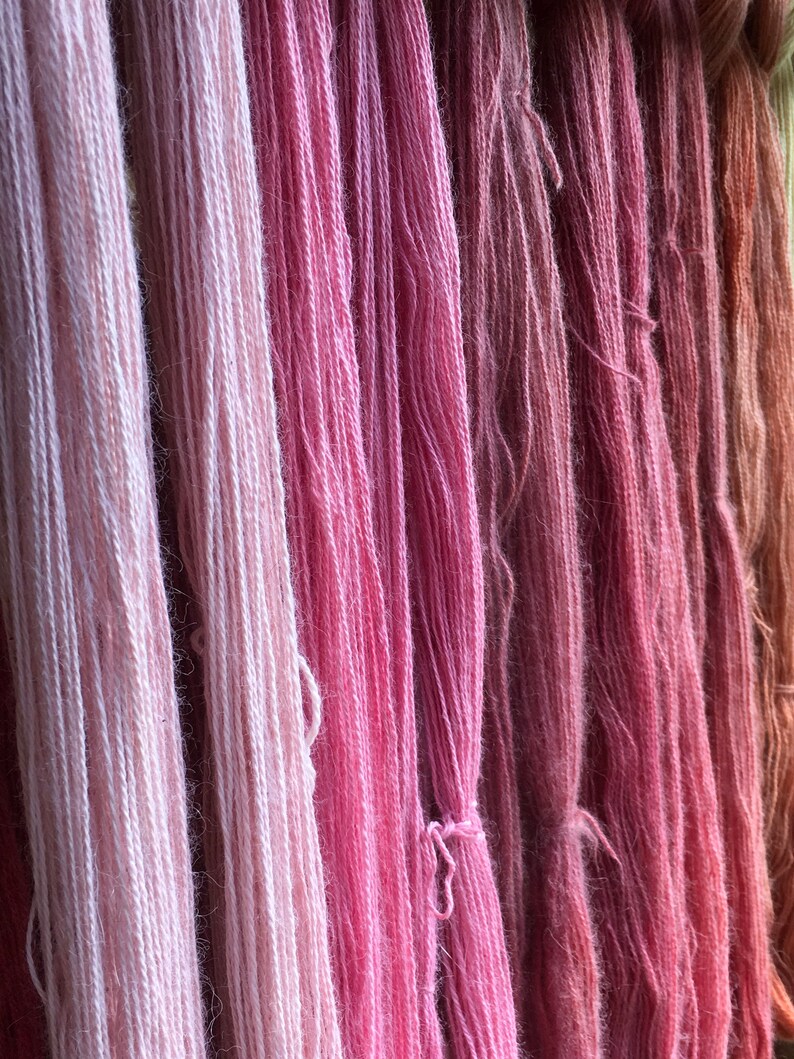 K&ouml;nnte beinhalten: Nahaufnahme von verschiedenfarbigen rosa Garnen, die vertikal h&auml;ngen. Die Farben reichen von hellrosa bis zu tiefem Ros&eacute; und erzeugen einen Farbverlauf. Die Textur des Garns ist sichtbar, was auf eine weiche, nat&uuml;rliche Faser hindeutet. Ideal f&uuml;r Strick- und H&auml;kelprojekte.