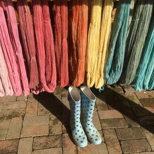 K&ouml;nnte beinhalten: Verschiedenfarbige Garne in Rosa, Rot, Gelb und Blau h&auml;ngen zum Trocknen. Hellblaue Gummistiefel mit schwarzen Punkten stehen vor den Garnen auf einem Ziegelboden.