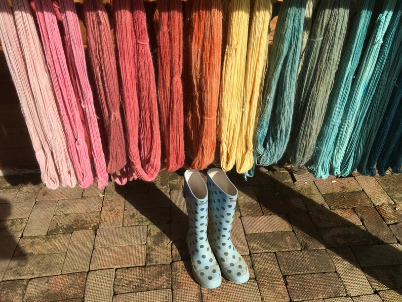 K&ouml;nnte beinhalten: Verschiedenfarbige Garne in Rosa, Rot, Gelb und Blau h&auml;ngen zum Trocknen. Hellblaue Gummistiefel mit schwarzen Punkten stehen vor den Garnen auf einem Ziegelboden.