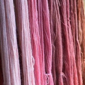 K&ouml;nnte beinhalten: Nahaufnahme von mehreren Garnstr&auml;ngen in verschiedenen Rosa- und Pfirsicht&ouml;nen. Das Garn ist vertikal angeordnet und erzeugt eine strukturierte und farbenfrohe Darstellung. Die Farben reichen von hellem Rosa bis zu tieferen Rosen- und Korallt&ouml;nen, was auf einen nat&uuml;rlichen F&auml;rbeprozess hindeutet.
