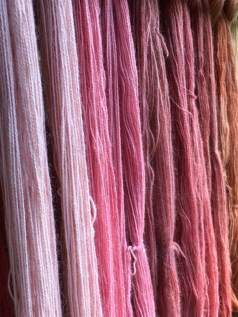 K&ouml;nnte beinhalten: Nahaufnahme von mehreren Garnstr&auml;ngen in verschiedenen Rosa- und Pfirsicht&ouml;nen. Das Garn ist vertikal angeordnet und erzeugt eine strukturierte und farbenfrohe Darstellung. Die Farben reichen von hellem Rosa bis zu tieferen Rosen- und Korallt&ouml;nen, was auf einen nat&uuml;rlichen F&auml;rbeprozess hindeutet.