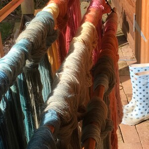 K&ouml;nnte beinhalten: Bunte Wollkn&auml;uel in Blau-, Gr&uuml;n-, Rosa- und Oranget&ouml;nen werden auf einem Holzgestell pr&auml;sentiert. Die Wolle ist in einem Farbverlauf angeordnet, wobei jedes Kn&auml;uel eine Mischung aus Farben zeigt. Ein Paar gepunktete Gummistiefel ist im Hintergrund zu sehen.