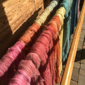 K&ouml;nnte beinhalten: Ein Holzgestell zeigt mehrere Str&auml;nge farbiger Wolle, die in einem Farbverlauf gef&auml;rbt wurden. Die Wolle reicht von Rosa und Orange bis hin zu Gelb und Blau, wobei jeder Strang um einen Holzd&uuml;bel gewickelt ist. Die Szene wird von Sonnenlicht beleuchtet.