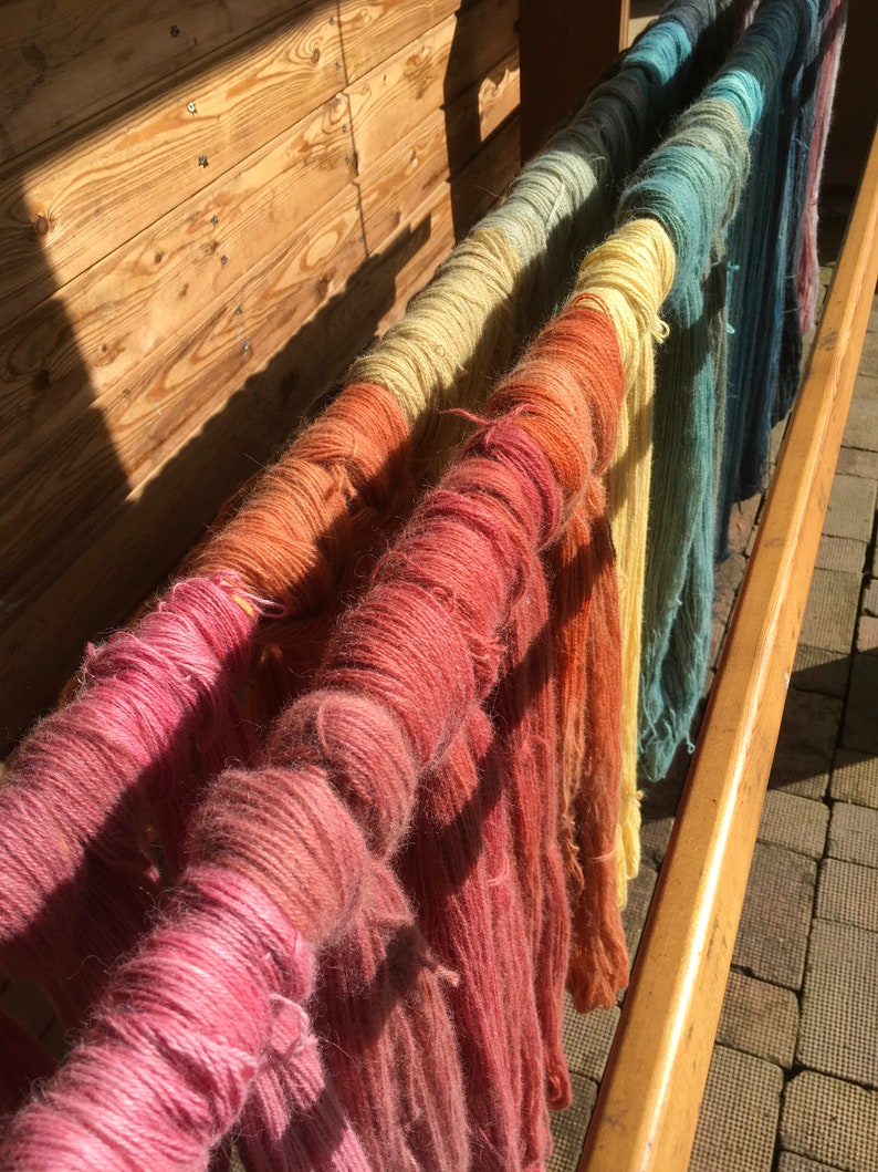 K&ouml;nnte beinhalten: Ein Holzgestell zeigt mehrere Str&auml;nge farbiger Wolle, die in einem Farbverlauf gef&auml;rbt wurden. Die Wolle reicht von Rosa und Orange bis hin zu Gelb und Blau, wobei jeder Strang um einen Holzd&uuml;bel gewickelt ist. Die Szene wird von Sonnenlicht beleuchtet.