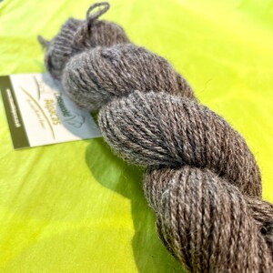 Puede incluir: Un ovillo de lana de alpaca marr&oacute;n. La lana est&aacute; retorcida en una bobina suelta y est&aacute; sobre una superficie verde. La etiqueta dice "Peruvian Alpaca" con un peque&ntilde;o icono de alpaca.