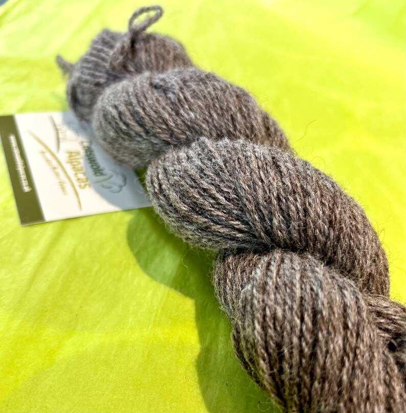 Puede incluir: Un ovillo de lana de alpaca marr&oacute;n. La lana est&aacute; retorcida en una bobina suelta y est&aacute; sobre una superficie verde. La etiqueta dice "Peruvian Alpaca" con un peque&ntilde;o icono de alpaca.