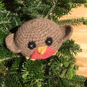 May include: A brown crocheted robin ornament with black eyes, a yellow beak, and a red breast. The ornament is nestled in a green fir tree.