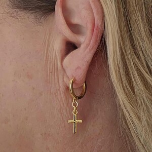 Op de afbeelding: Een goudkleurige oorring met een hangende kruisvormige hanger. De oorring is bevestigd aan een oor. Het kruis is een eenvoudig, klassiek ontwerp. Het is een religieus symbool en een sieraad.