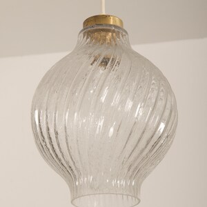 May include: Clear glass pendant light with a textured, swirled design. The light fixture has a bulbous shape with a brass-colored top and a white cord for hanging. The glass has a frosted appearance.