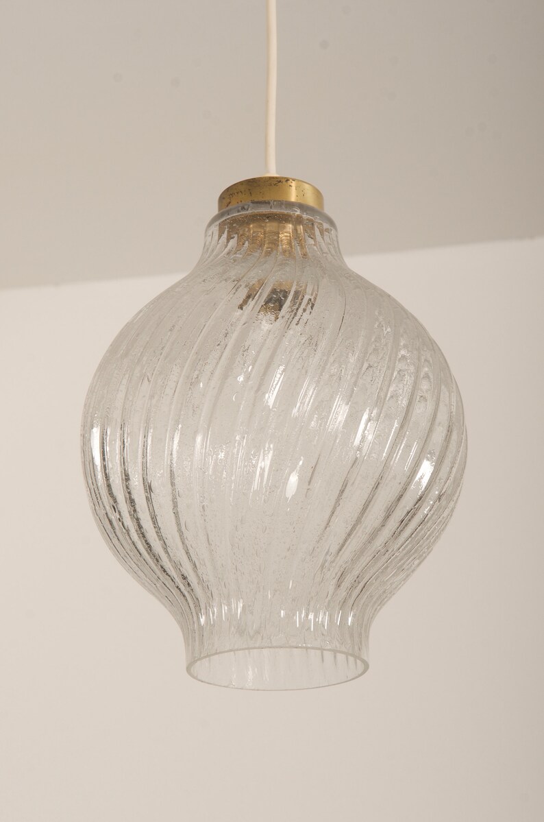 May include: Clear glass pendant light with a textured, swirled design. The light fixture has a bulbous shape with a brass-colored top and a white cord for hanging. The glass has a frosted appearance.