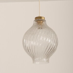 May include: Clear glass pendant light with a textured, swirling design. The light fixture has a bulbous shape and a brass-colored top. A white cord suspends the light from the ceiling. The light fixture is a decorative element for home decor.
