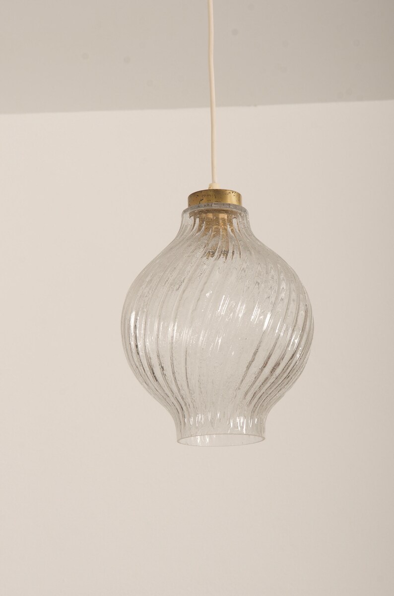 May include: Clear glass pendant light with a textured, swirling design. The light fixture has a bulbous shape and a brass-colored top. A white cord suspends the light from the ceiling. The light fixture is a decorative element for home decor.