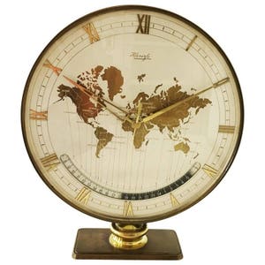 Puede incluir: Un reloj de mesa dorado con un diseño de mapa del mundo. La esfera del reloj presenta números romanos y una aguja de hora y minutos dorada. El reloj está sobre una base marrón.