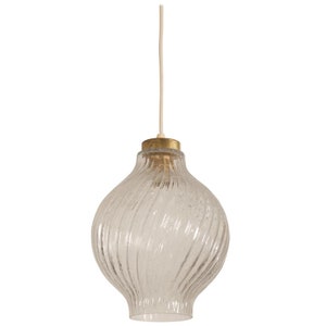 May include: Clear glass pendant light with a textured, swirled design. The light fixture has a bulbous shape and a brass-colored metal cap. A white cord suspends the light from above, creating a vintage aesthetic.