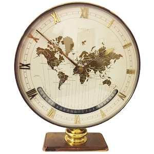 Puede incluir: Un reloj de mapa del mundo de tono dorado con marcadores de hora en números romanos y un diseño de mapa del mundo en blanco y negro. El reloj tiene una base de latón y un soporte de tono dorado.