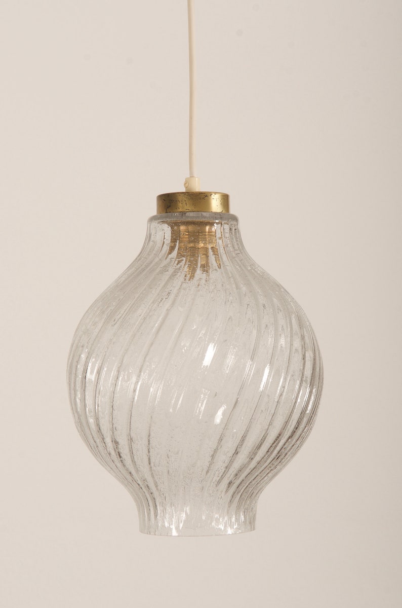 May include: Clear glass pendant light with a textured, swirled design. The light fixture has a brass-colored top and a white cord for hanging. This vintage-style light fixture is suitable for a kitchen or dining room.