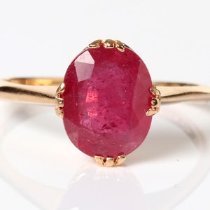 May include: A gold ring with a large, oval-shaped ruby gemstone. The ruby is a deep red color and has a faceted cut.