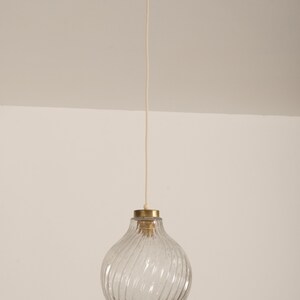 May include: A clear glass pendant light with a textured, swirled design. The light fixture has a brass-colored top and a white ceiling mount. The cord is a light beige color.