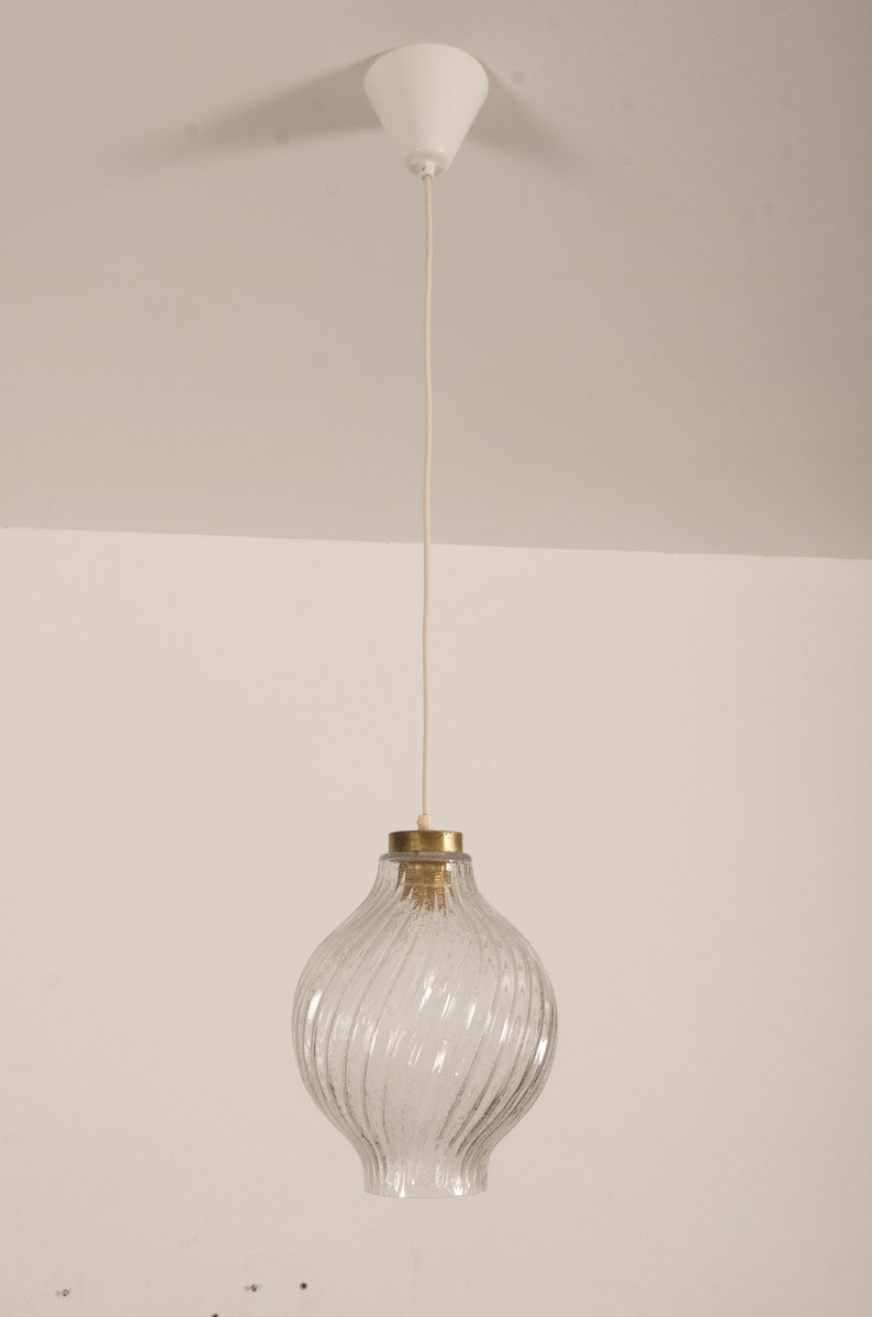May include: A clear glass pendant light with a textured, swirled design. The light fixture has a brass-colored top and a white ceiling mount. The cord is a light beige color.