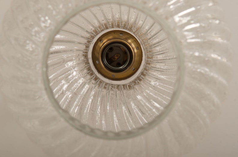 May include: A clear glass light fixture with a textured, swirled design. The fixture has a brass-colored socket and a white base. The light fixture is designed to be mounted on a ceiling.