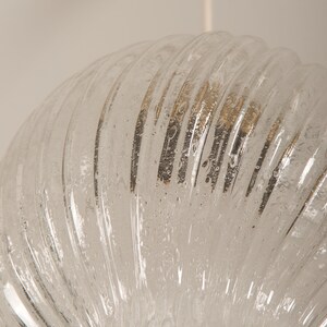 May include: Clear glass pendant light fixture with a ribbed, semi-circular shade. The shade has a textured surface with small bubbles and a white cord. The light fixture is suspended from the ceiling.
