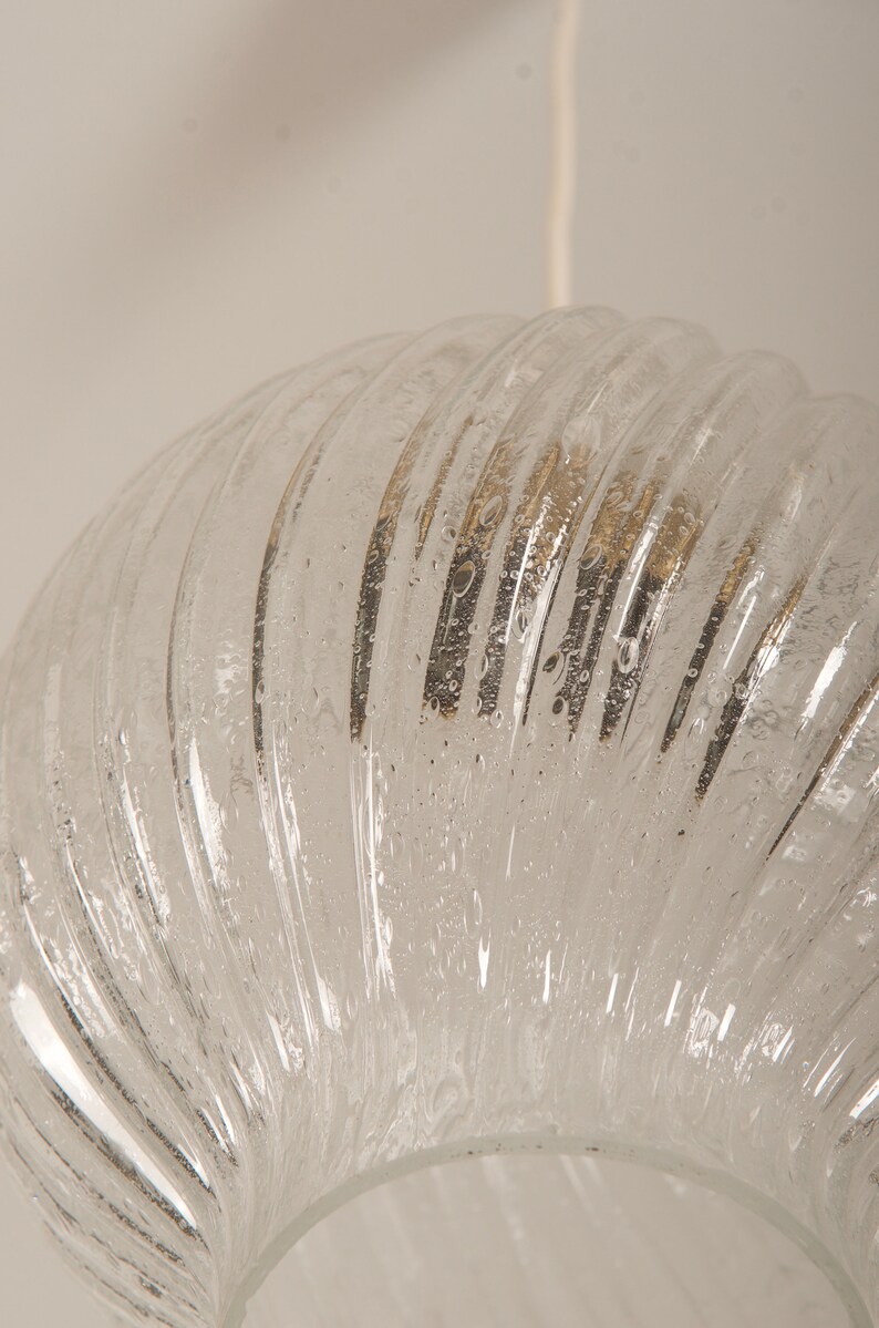 May include: Clear glass pendant light fixture with a ribbed, semi-circular shade. The shade has a textured surface with small bubbles and a white cord. The light fixture is suspended from the ceiling.
