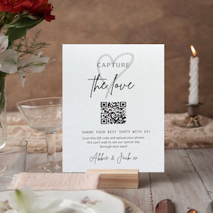 以下が含まれることがあります： 「CAPTURE the love」の文字とQRコードが書かれた白いカード。下には「SHARE YOUR BEST SHOTS WITH US!」と「Abbie & Jack xx」と書かれています。カードは木のブロックで支えられ、テーブルにはキャンドルと花が飾られています。