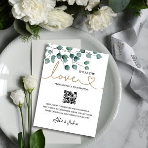 以下が含まれることがあります： 白い招待状カード。金色の筆記体で「love」と書かれ、水彩画のユーカリの葉で飾られています。カードにはQRコードと、ゲストに写真の共有を促すテキストが含まれています。カードは白いお皿の上にあり、花が添えられています。