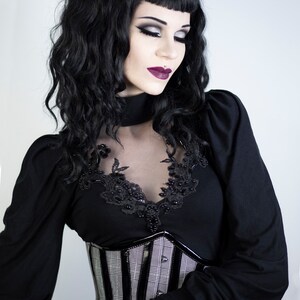 May include: A woman in a black gothic-style outfit. She wears a black top with lace and bead detailing, a plaid corset, and long sleeves. Her dark hair is styled with bangs, and she has dramatic makeup.