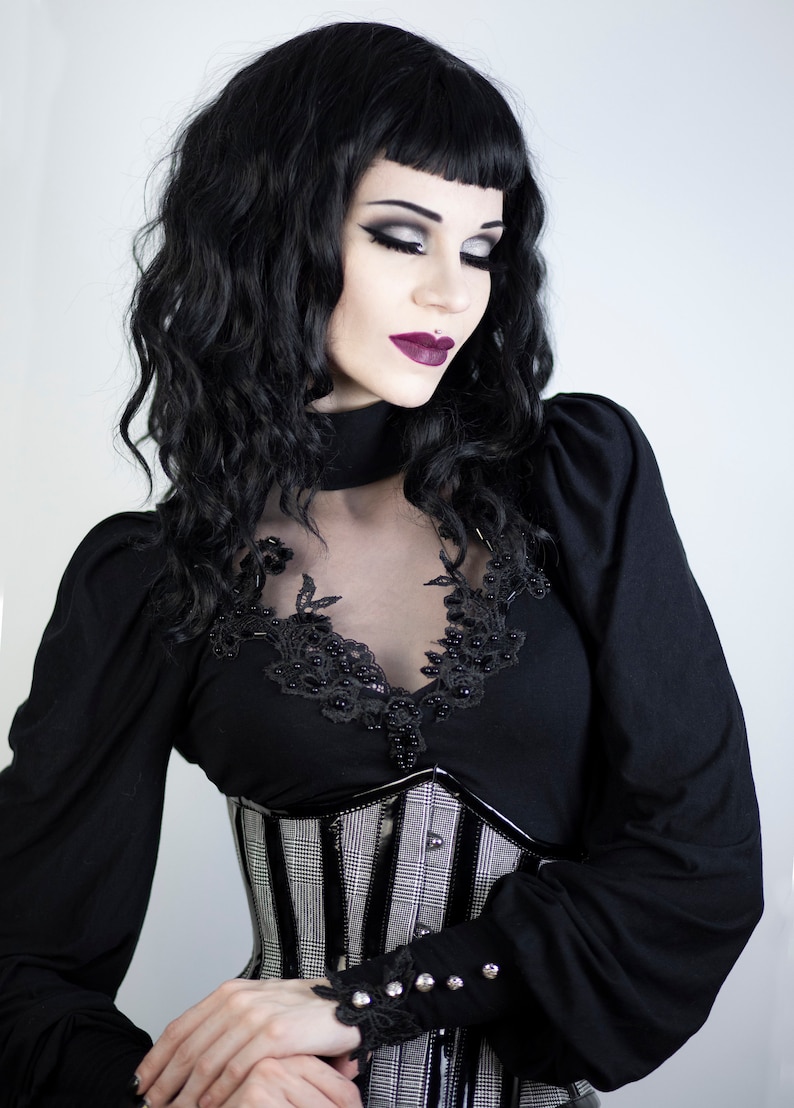 May include: A woman in a black gothic-style outfit. She wears a black top with lace and bead detailing, a plaid corset, and long sleeves. Her dark hair is styled with bangs, and she has dramatic makeup.