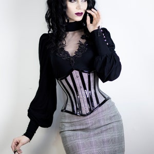 May include: A black top with long sleeves and a sheer neckline, paired with a striped corset and a gray plaid mini skirt. The corset has a glossy black trim. The outfit is styled in a gothic fashion.