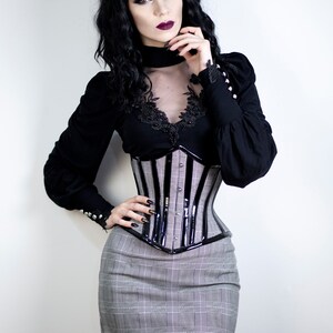 May include: A black long-sleeved top with lace detailing and a plaid mini skirt. The outfit includes a black and silver corset. The model has dark hair and is wearing dark lipstick and nail polish.