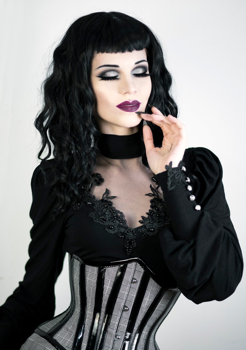 May include: A person wearing a black top with a sheer neckline, a black and white plaid corset, and dark makeup. The top has long sleeves with decorative buttons and lace cuffs. The person has dark hair and is wearing dark lipstick.