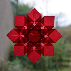 Op de afbeelding: Een rode papieren sneeuwvlokdecoratie met een geometrisch ontwerp. De sneeuwvlok is gemaakt van meerdere lagen papier, waardoor een driedimensionaal effect ontstaat.
