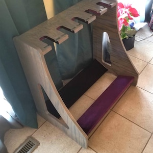 May include: A wooden guitar stand with a gray finish. The stand has slots for multiple guitars and features black and purple velvet-covered shelves. The stand is designed to hold several guitars and is made of wood.